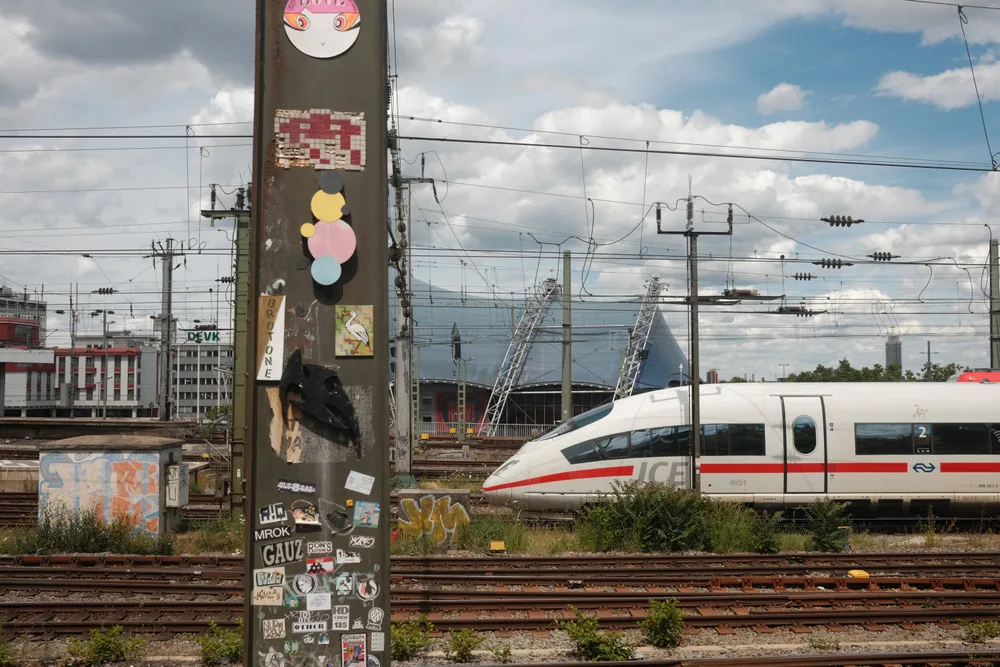 A white high-speed train with a red stripe among tracks and overhead wires, behind a sticker-covered pole
