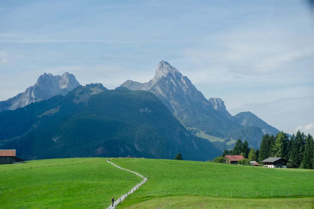 A winding path across a green meadow towards houses beneath rocky mountain peaks